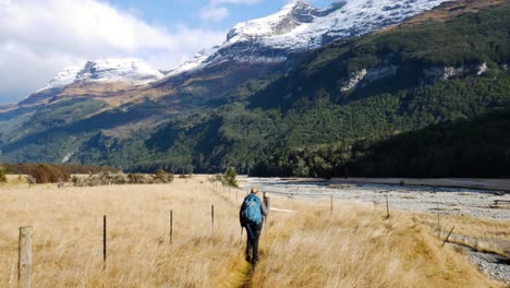 Vista-Trasera-De-Una-Mujer-Rubia-Con-Mochila-Y-Bastones-De-Senderismo-En-Pastos-Amarillos-Durante-El-Día-Soleado---Hermosa-Cordillera-Con-Picos-Nevados-En-El-Fondo