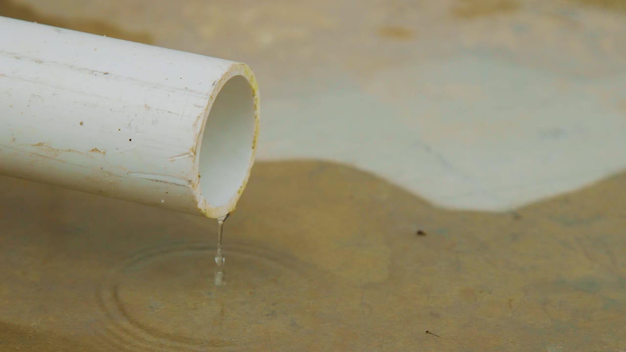 Water slowly dripping on a concrete floor from a white PVC pipe making a puddle - Close up