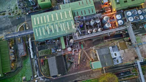 Rotating-Overhead-drone-aerial-footage-of-a-large-industrial-plant-showing-pipework-structures,-buildings,-cooling-towers,-steam,-and-work-vehicles