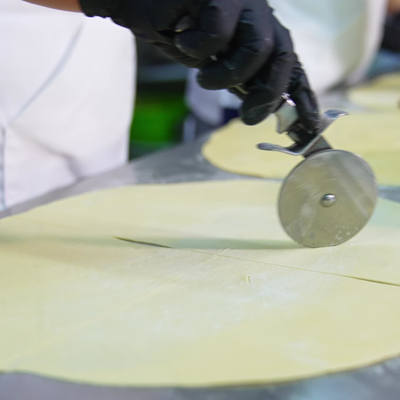 Baker uses pizza knife for cutting dough rolled on the table. Close up. Other workers put the stuffing on the pastry in blur. Croissants making