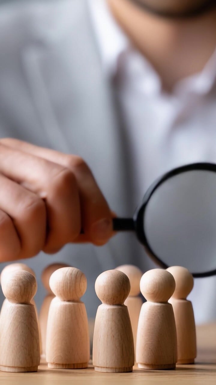 Analyzing Potential Candidates: A Business Professional Using a Magnifying Glass to Inspect Wooden Figures Representing Team Members in a Corporate Setting