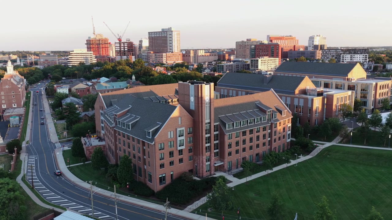 Un'auto percorre una strada curva oltre un edificio dormitorio in mattoni a New Brunswick, New Jersey; lo skyline del centro e le gru edili si ergono oltre prati verdi alla luce del tramonto. Vista aerea con drone