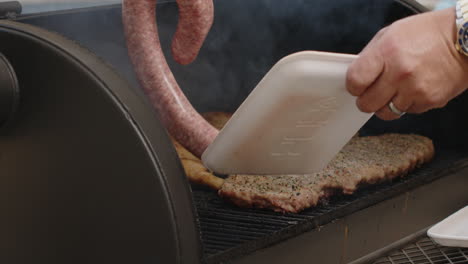 slow motion of tongs placing bbq sausage on a smoking grill pit with ribs and chicken thighs