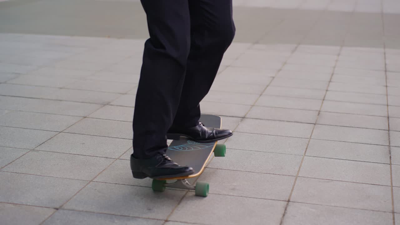 Carrellata sui piedi di un professionista in pantaloni neri e scarpe di cuoio che esegue acrobazie sullo skateboard mentre si reca in ufficio in una giornata di sole.