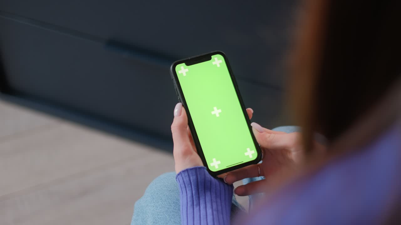 Young woman sitting on sofa at home and uses smartphone with green screen Mock-up. Holding in hand and looking on the screen. 4k