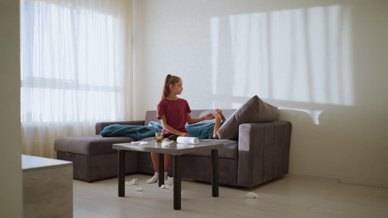 Caring girl hands thermometer to feverish brother to check his temperature, tissue littered around with a cozy couch setting and shadows from natural light casting on the wall