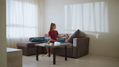 caring girl hands thermometer to feverish brother to check his temperature, tissue littered around with a cozy couch setting and shadows from natural light casting on the wall