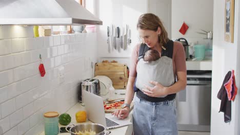 Video-of-caucasian-mother-with-newborn-baby-in-baby-carrier-using-laptop-in-kitchen