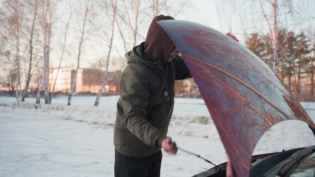 Young man in hooded winter jacket opens car bonnet and places support rod to secure hood in snowy outdoor area surrounded by leafless trees and distant buildings