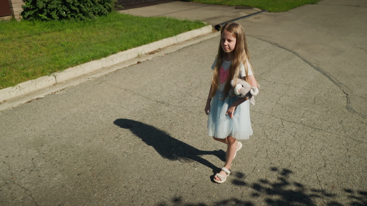 Little girl squinting under bright sunlight walks alone on cracked tarred road holding plush toy, with grass-lined pavement and house in background, casting soft shadow on textured asphalt surface