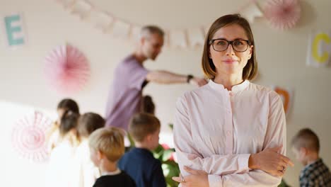 Porträt-Einer-Lehrerin-Mit-Brille-Und-Rosa-Hemd,-Die-Mit-Verschränkten-Armen-Vor-Der-Brust-Lächelt,-Vor-Dem-Hintergrund-Einer-Biologiestunde-Für-Vorschulkinder,-Zusammen-Mit-Einem-Männlichen-Lehrer-In-Einem-Lila-Hemd