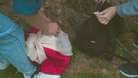 primer plano de mujeres con vaqueros y zapatillas de deporte recuperando mochilas del suelo del bosque, cerrándolas con las manos con cuidado sosteniendo bolsas, rodeadas de agujas de pino, hierba