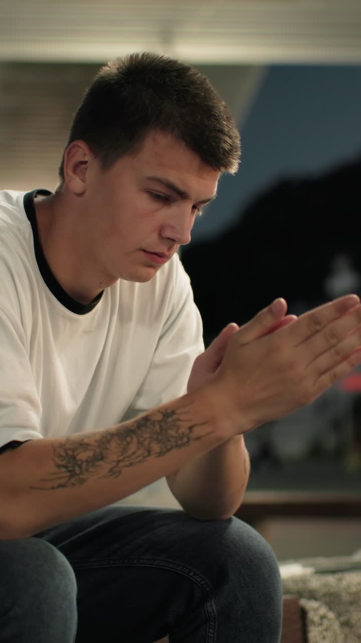 Reflective man seated on bench with tattooed hand at dusk, looking thoughtfully, city lights and urban structures in background at night