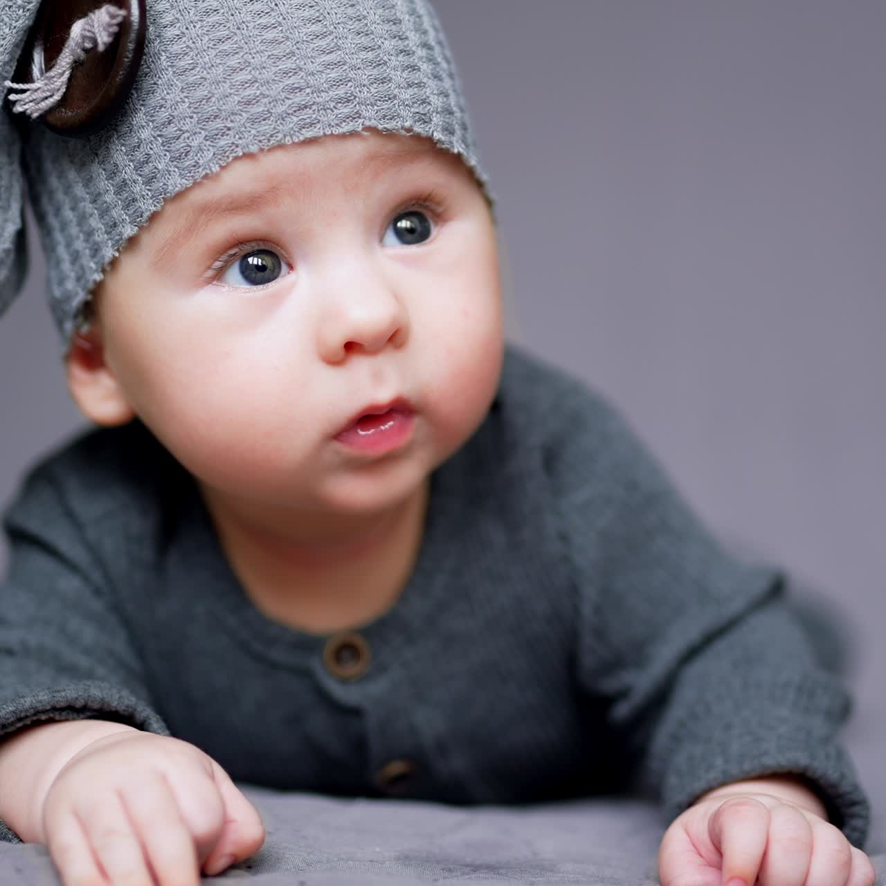 Tiny cute boy in funny grey hat lying on his belly. Lovely kid wearing grey clothes lying at grey backdrop. Close up