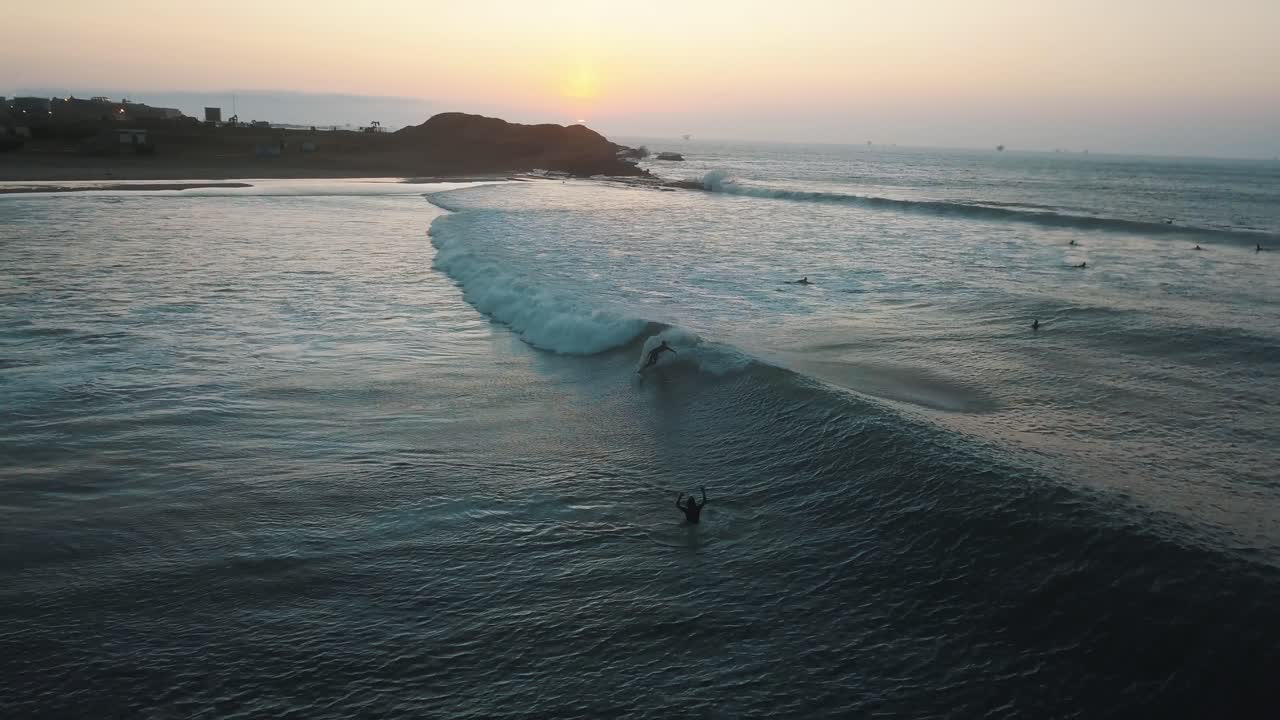 Aerial view of surfers catching waves, during sunset, at the coast of Talara, in Peru - tracking, drone shot