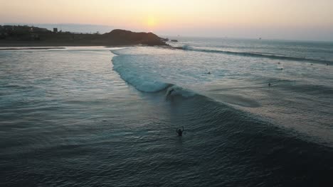 aerial view of surfers catching waves, during sunset, at the coast of talara, in peru - tracking, drone shot