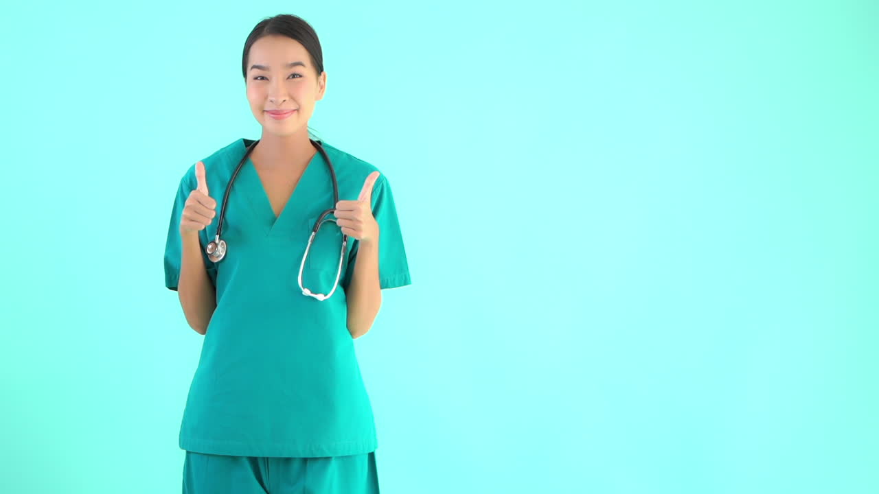 Young attractive Asian woman in scrubs smiles as she raises her hands with a thumbs up. Copy space
