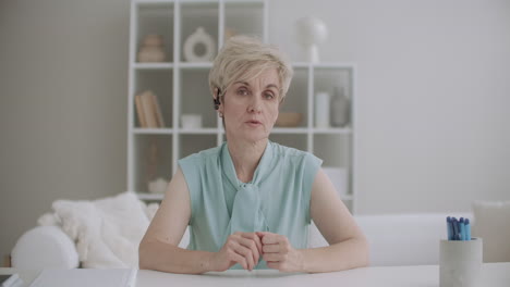 aged woman is lecturing online sitting at home at table and looking at camera video call or conference concept