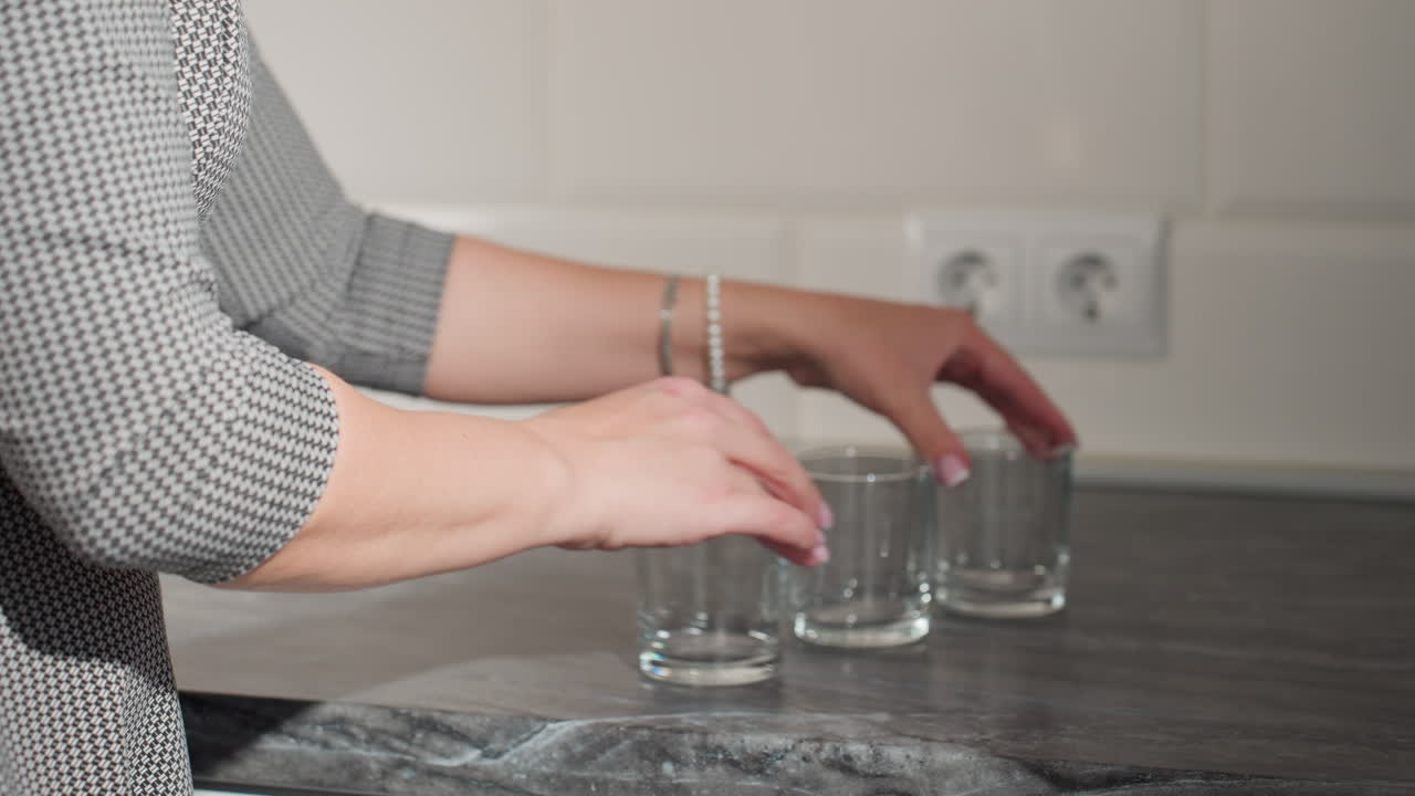 Hand view of girl in checkered gown playfully moving cup on kitchen countertop, engaging in playful game, electric outlet visible on wall, lively kitchen atmosphere filled with fun and energy