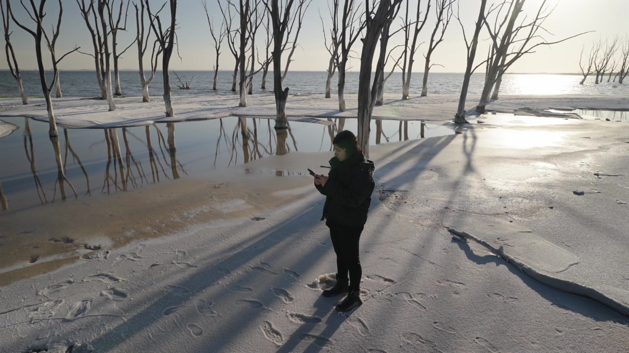 Donna che scrive messaggi al Lago Epecuén ad Adolfo Alsina, Provincia di Buenos Aires, Argentina, in piedi tra alberi allagati e acqua ipersalina riflettente sotto la luce bassa del sole, inquadratura statica.