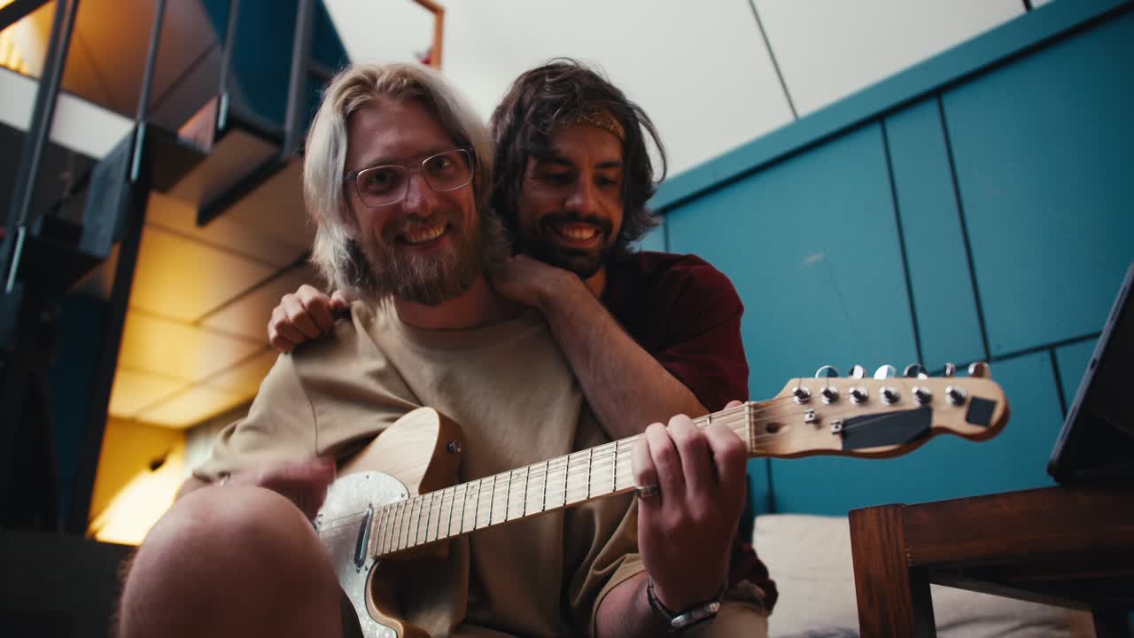Two guys friend spending time together guy brunette hugging guy blonde with a beard in glasses who plays the electric guitar in a cozy room