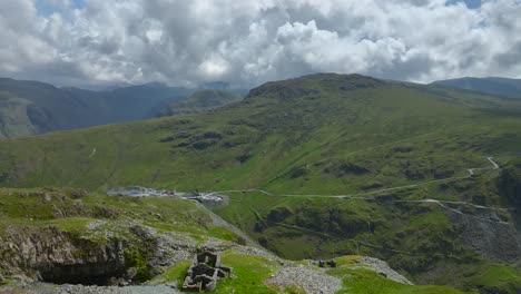 Derelict-slate-mine-crumbling-buildings-on-mountainside-with-drone-flight-over-towards-deep-valley-slate-mine-and-distant-mountains-under-overcast-sky