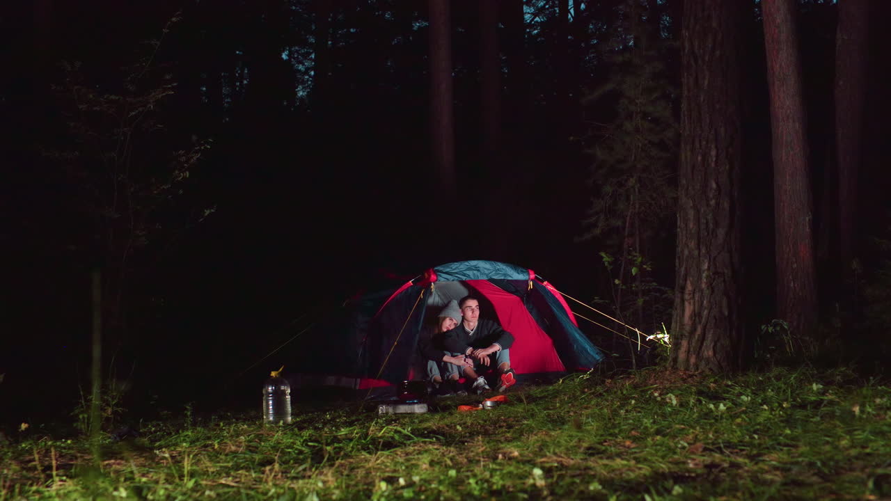 Lady rests head on boyfriend shoulder while seated inside tent during peaceful night camping in forest, surrounded by trees and soft glowing light