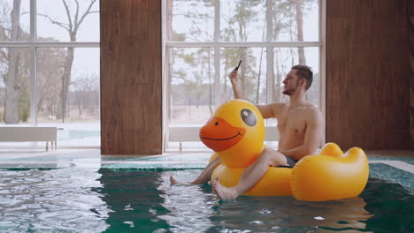 man is floating on inflatable circle in swimming pool in hotel and calling by video in smartphone