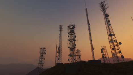 Cámara-Panorámica-Que-Muestra-Un-Complejo-De-Antenas.-El-Mástil-De-Radio-Sostiene-Antenas-Para-Telecomunicaciones,-Radiodifusión-Y-Televisión.-Estructuras-Altas-Con-Montañas-Y-Un-Cielo-Al-Atardecer-De-Fondo.-4k