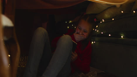 caucasian girl smiling while using smartphone under blanket fort during christmas at home