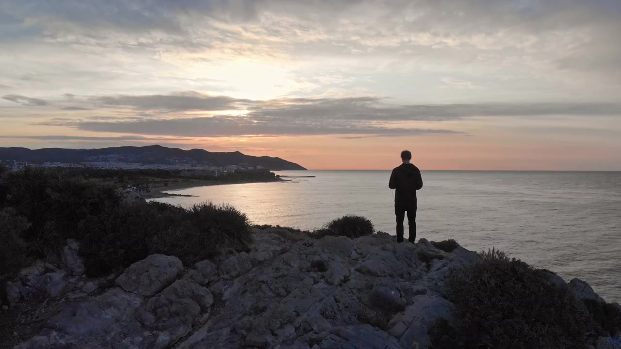 Un drone vola oltre la sagoma di un uomo in piedi su una scogliera durante l'alba sul mare