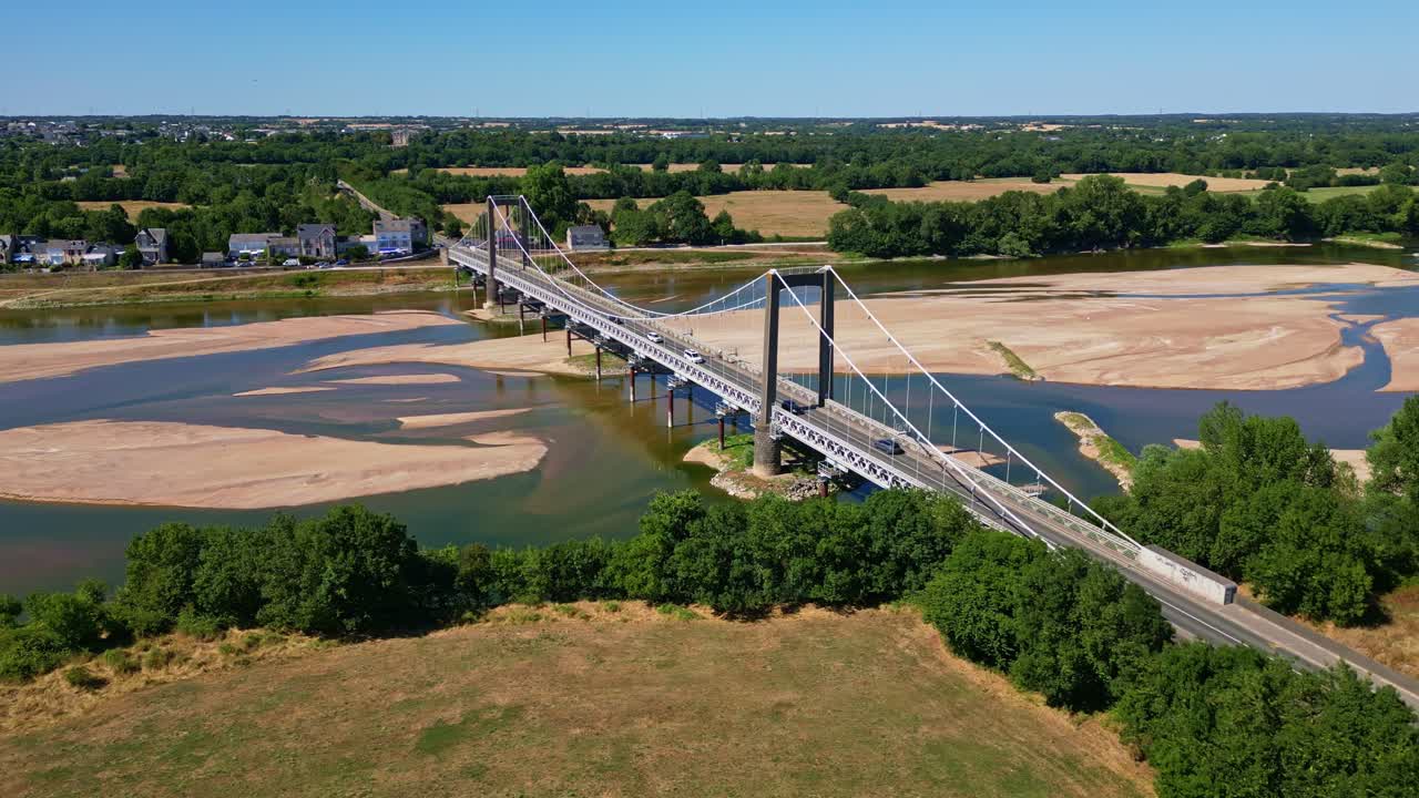 Drone lateral shot of Varades suspension bridge crossing Loire River, showing sandbanks, greenery, and vehicles in motion - France