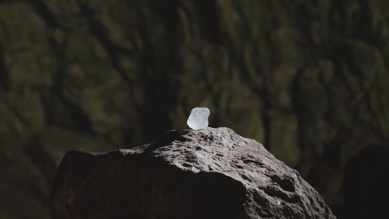 Cinematic shot of a luminous raw crystal resting on a rock pedestal in a dark cave. Mysterious smoke drifts by, creating a magical or sacred fantasy atmosphere