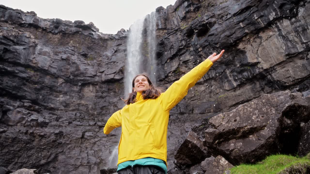 una mujer con una chaqueta amarilla se para alegremente frente a una cascada alta al aire libre