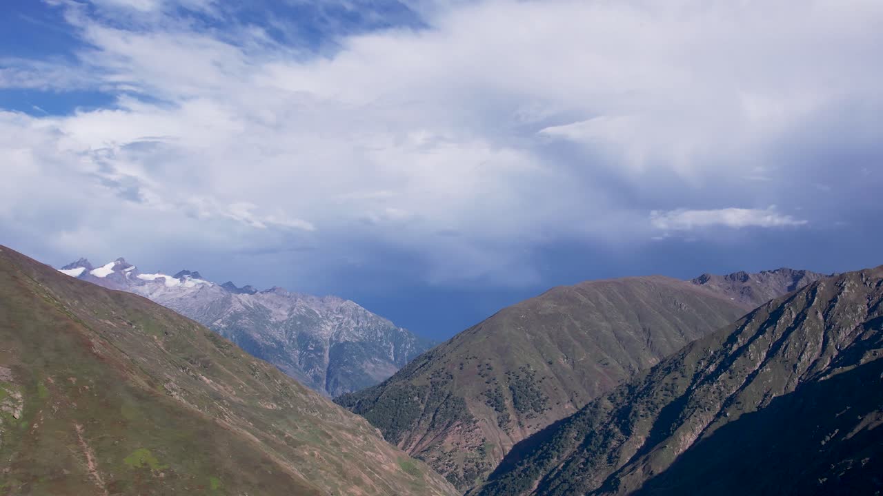 Layered Himalayan ridges with snow-capped peaks in the distance beneath a textured, cloudy sky