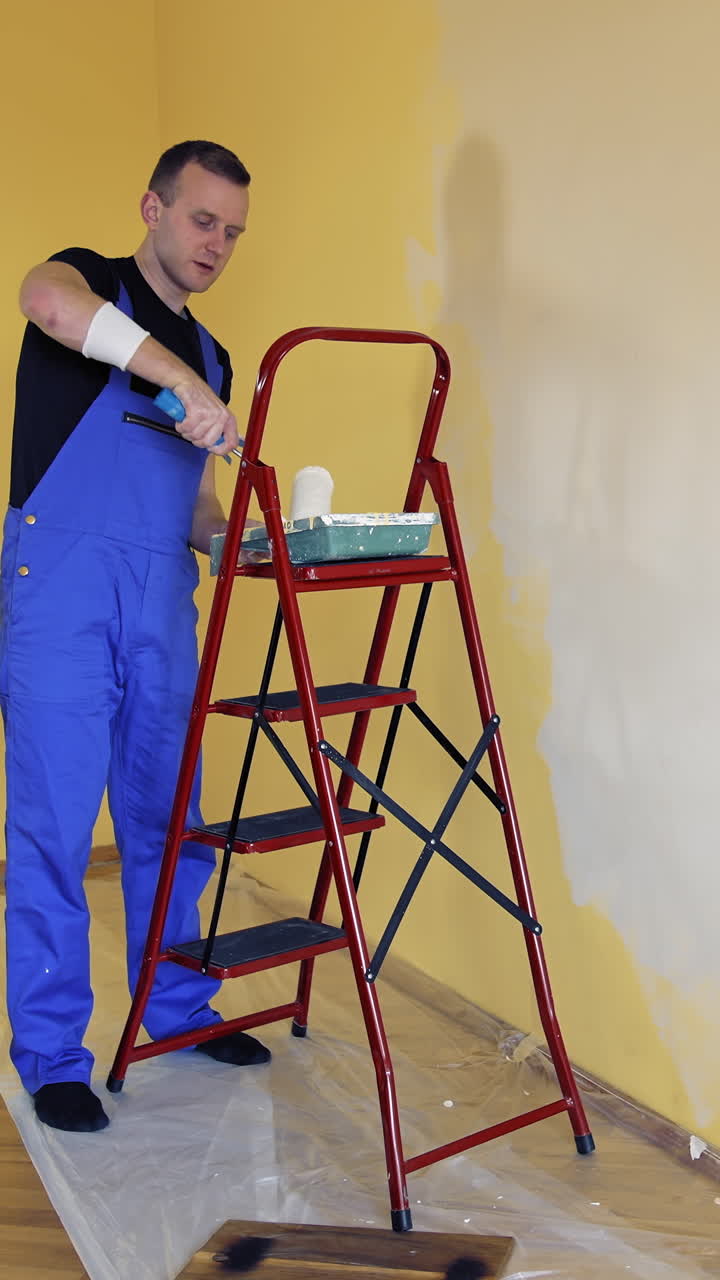 Young man in overalls renovating room in the house. Painter worker using a paint roller doing makeover of a room. Room decoration with new color. Vertical video