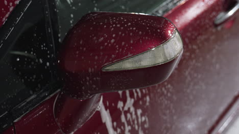 detailed close up of red car side mirror covered in soapy droplets and water drops during washing process showing clear reflections, textured surface, and dynamic water patterns inside indoor garage