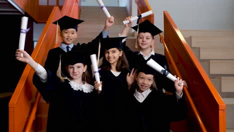 Group-Of-Happy--Preschool-Students-In-Cap-And-Gown-Standing-On-Stairs,-Looking-At-The-Camera-And-Showing-Their-Graduation-Diplomas