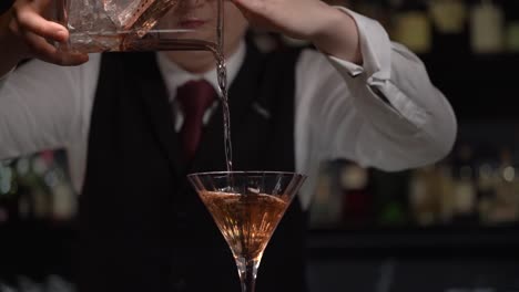 a young barmaid is elegantly pouring the slightly diluted chilled martini into a cocktail glass using a copper strainer and cristal stirring glass.