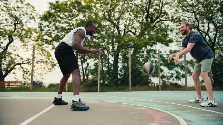 Eine schwarze Person in einem weißen T-Shirt und ein rothaariger Mann in einem grauen T-Shirt werfen sich auf einem Basketballplatz einen Ball zu. Der rothaarige Mann versucht, den Ball in den Basketballkorb zu werfen