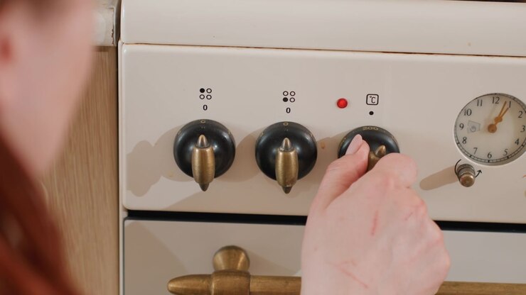 Vista aproximada de uma mão girando o botão de controle do forno em um fogão de esmalte branco, com três mostradores pretos, ponteiros metálicos e a superfície do eletrodoméstico de cozinha sugerindo ajuste ativo de temperatura, para uso doméstico.