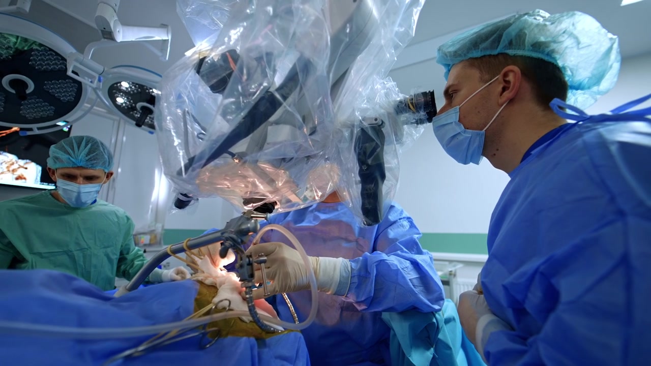 Kyiv, Ukraine, 5 May 2026: Neurosurgeons sharing binocular microscope during collaborative brain surgery. Both lean into draped eyepieces, scrub nurse at instrument table, monitor glowing behind.