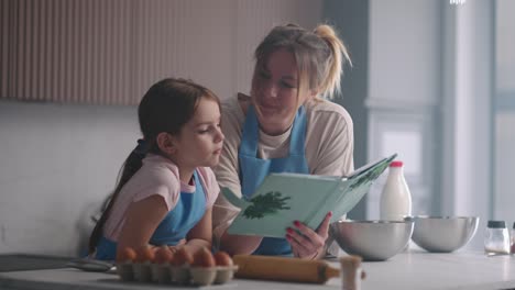 woman and her little daughter are reading cookbook kitchen of house in sunday, mother is teaching her child to cook