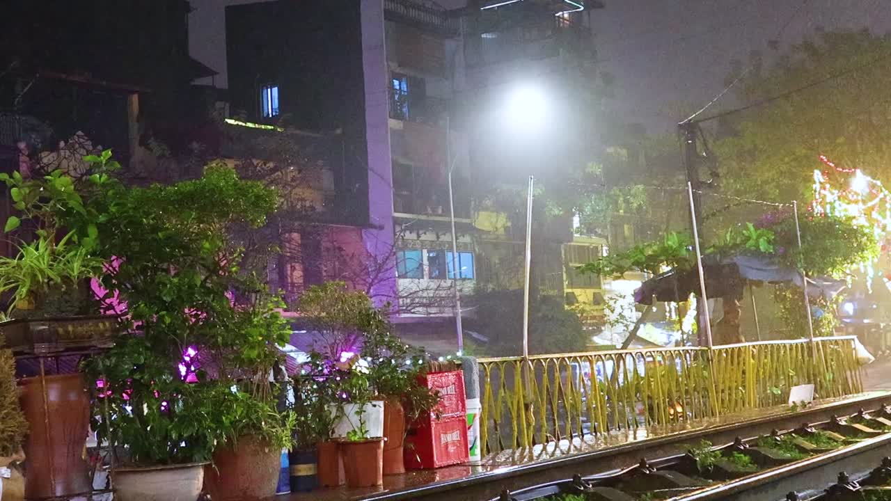 A train moves through a brightly lit street lined with lush plants and vibrant urban scenery at night.