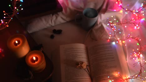 cozy background of an open book on a bed, next to a mug and surrounded by colorful christmas lights