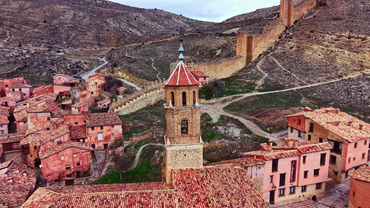 Pueblo histórico con tejados rojos y un campanario rodeado de montañas en España durante el día.