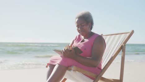 Happy-senior-african-american-woman-sitting-on-deck-chair-and-reading-book-at-beach,-in-slow-motion