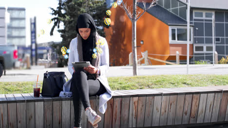 Animation-of-mixed-race-woman-wearing-hijab-in-street-using-tablet-computer-with-floating-emoji-symb