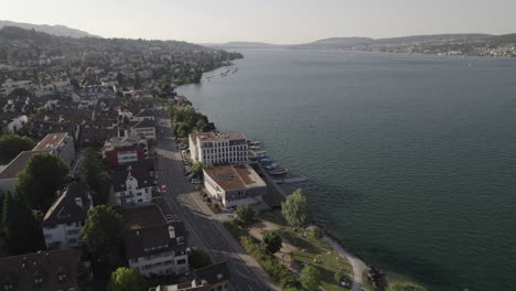 Drone-shot-flying-near-the-city-of-Thalwil-at-the-Zurich-Lake-in-Switzerland-on-a-sunny-day-with-water-and-boats-around-the-shore-and-white-houses-in-the-city-LOG