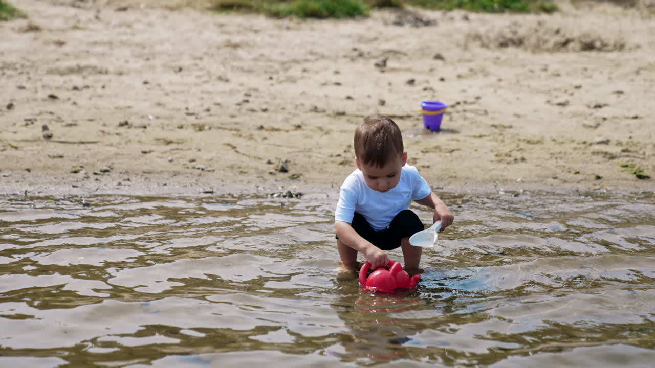Charming baby boy picks some water from the river. Kid walks by the water holding a watering can and a shovel.